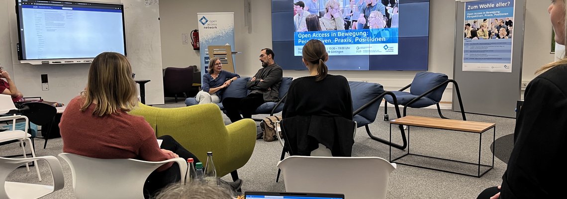 Diskussionsrunde moderiert von Margo Bargheer, rechts neben ihr auf dem Sofa: Andrew S. Gross vom New American Studies Journal Das Bild zeigt einen Vortragssaal von hinten mit Blick auf ein Sofa. Auf dem Sofa sitzen die Moderatorin Margo Bargheer und rechts neben ihr Andrew S. Gross vom New American Studies Journal.